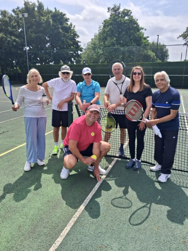 Seven members of the tennis group stand together at the net with their rackets, smiling at the camera.