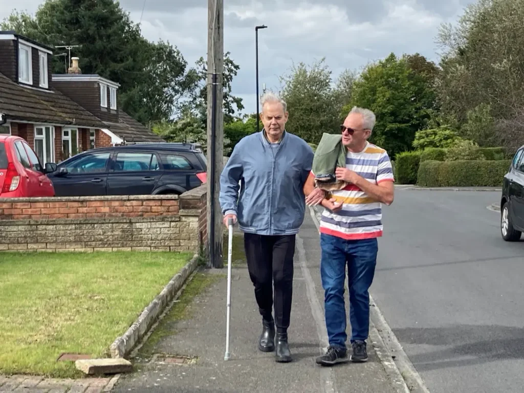 Two men walk along a residential street, one holding the other's arm to guide him as he uses a white cane.