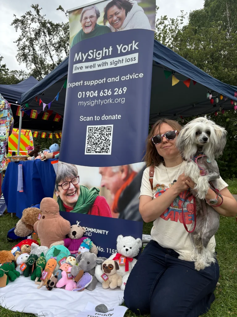 A person kneels beside a MySight York display at an outdoor community fair, holding a small dog. Behind them is a large MySight York banner featuring information about support services, a website address, phone number and a QR code for donations.