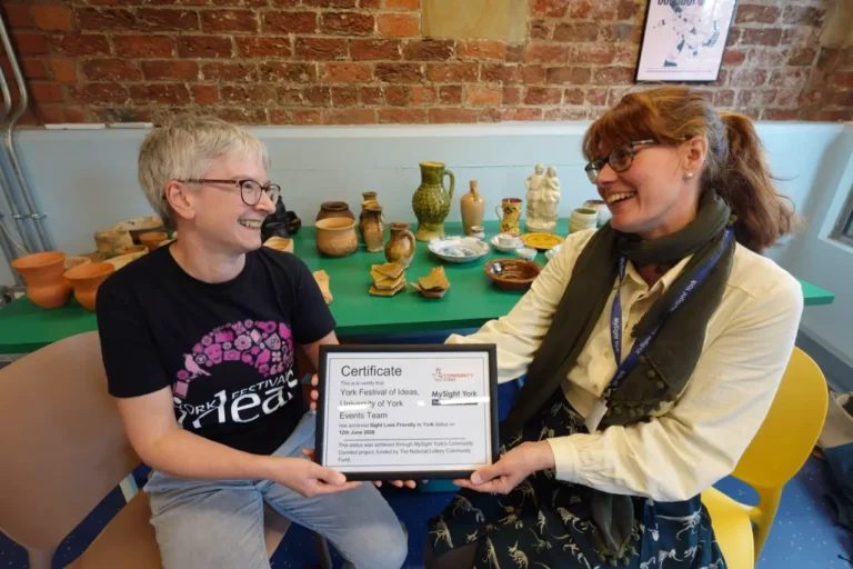 Two women laugh together as they hold a framed certificate awarding Sight Loss Friendly in York status to the York Festival of Ideas events team.