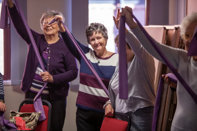 Several people take part in an indoor exercise class, standing in a circle and stretching purple resistance bands above and around their bodies.