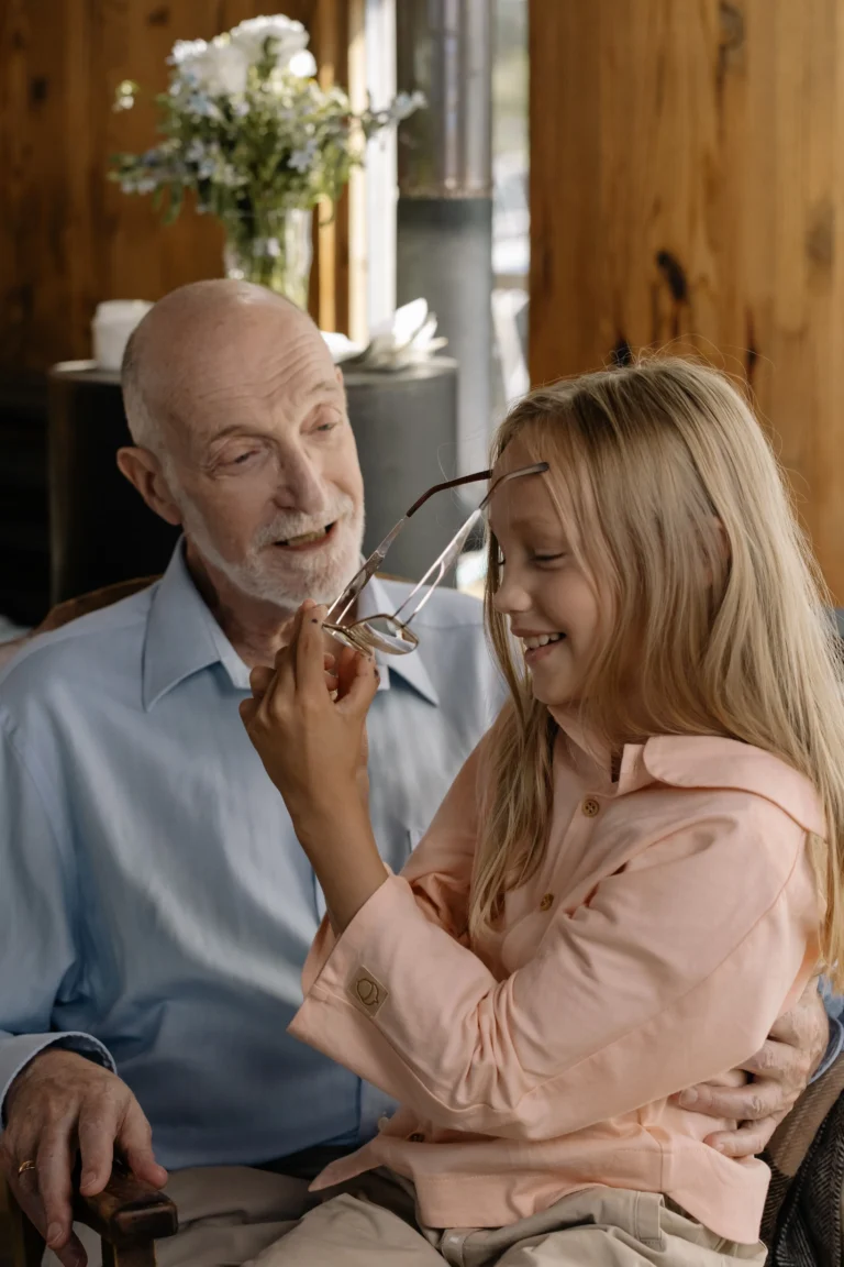 An older adult and a child sit together indoors in a warmly lit room with wooden walls. The child is holding a pair of eyeglasses up for a closer look while seated on the older adult’s lap.