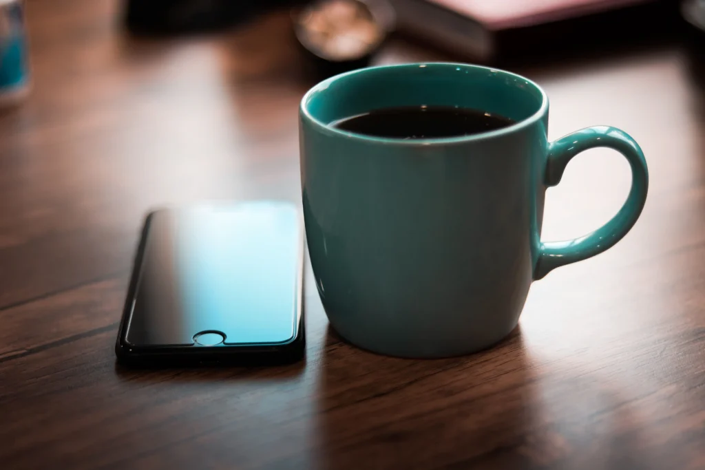 A teal ceramic mug filled with black coffee sits on a wooden desk beside a smartphone with a dark, reflective screen. The background is softly blurred, creating a warm, cosy atmosphere.