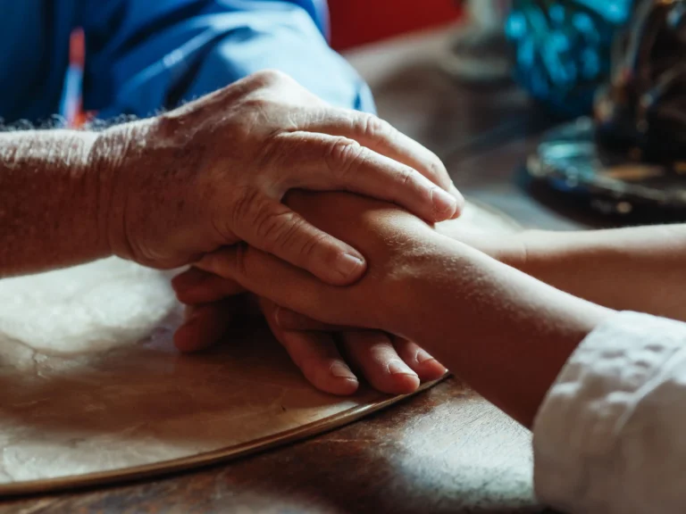 Close-up of two hands resting on a wooden table, with one hand gently placed over the other in a supportive gesture.