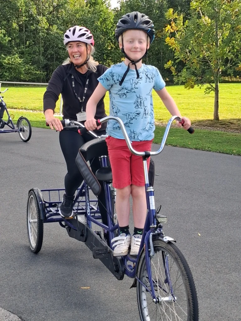 Two people wearing cycle helmets ride a tandem tricycle on a paved path. One person sits at the rear while a child stands at the front handlebars, with trees and grass in the background.