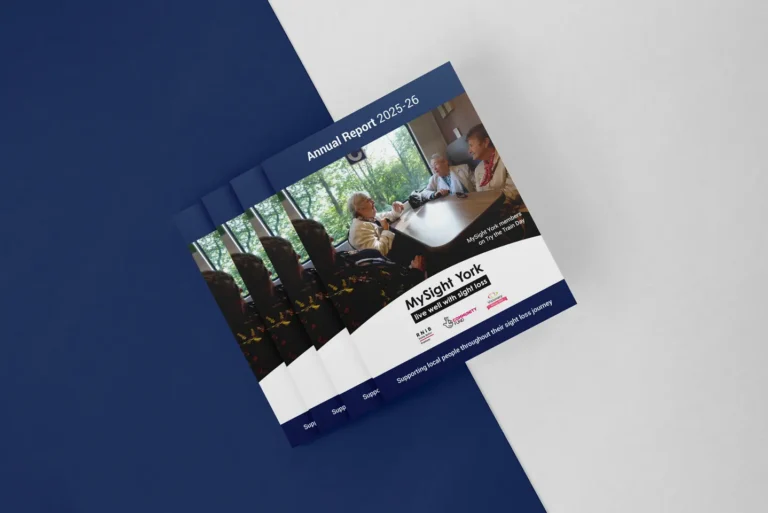 A stack of MySight York Annual Report 2025–26 booklets on a navy and grey background. The cover shows three members chatting around a table on a Try the Train Day, with the MySight York logo, strapline "live well with sight loss", and funder logos below.