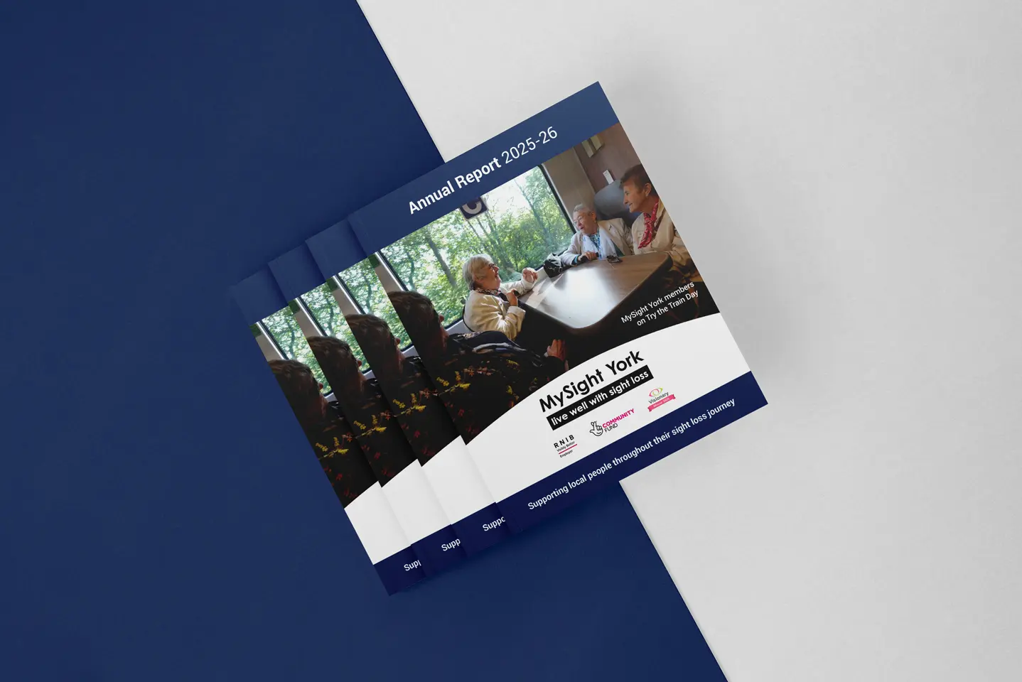 A stack of MySight York Annual Report 2025–26 booklets on a navy and grey background. The cover shows three members chatting around a table on a Try the Train Day, with the MySight York logo, strapline "live well with sight loss", and funder logos below.