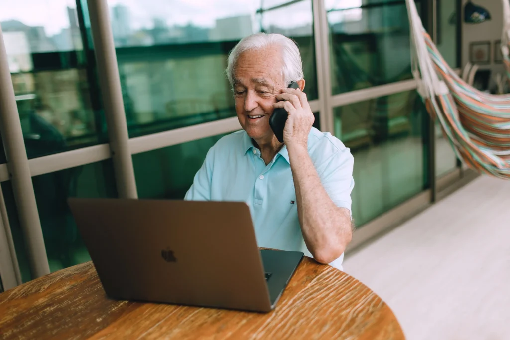 Person using a laptop while talking on a smartphone, sat at a table.
