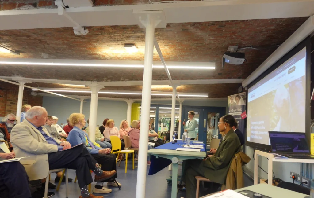 Members, volunteers and supporters attend the MySight York Annual General Meeting. Attendees are seated facing a speaker (Bethan, a Trustee at MySight York) and presentation screen which is displaying our new website, with some Trustees seated at a table at the front of the room.