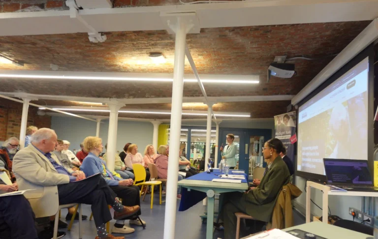 Members, volunteers and supporters attend the MySight York Annual General Meeting. Attendees are seated facing a speaker (Bethan, a Trustee at MySight York) and presentation screen which is displaying our new website, with some Trustees seated at a table at the front of the room.